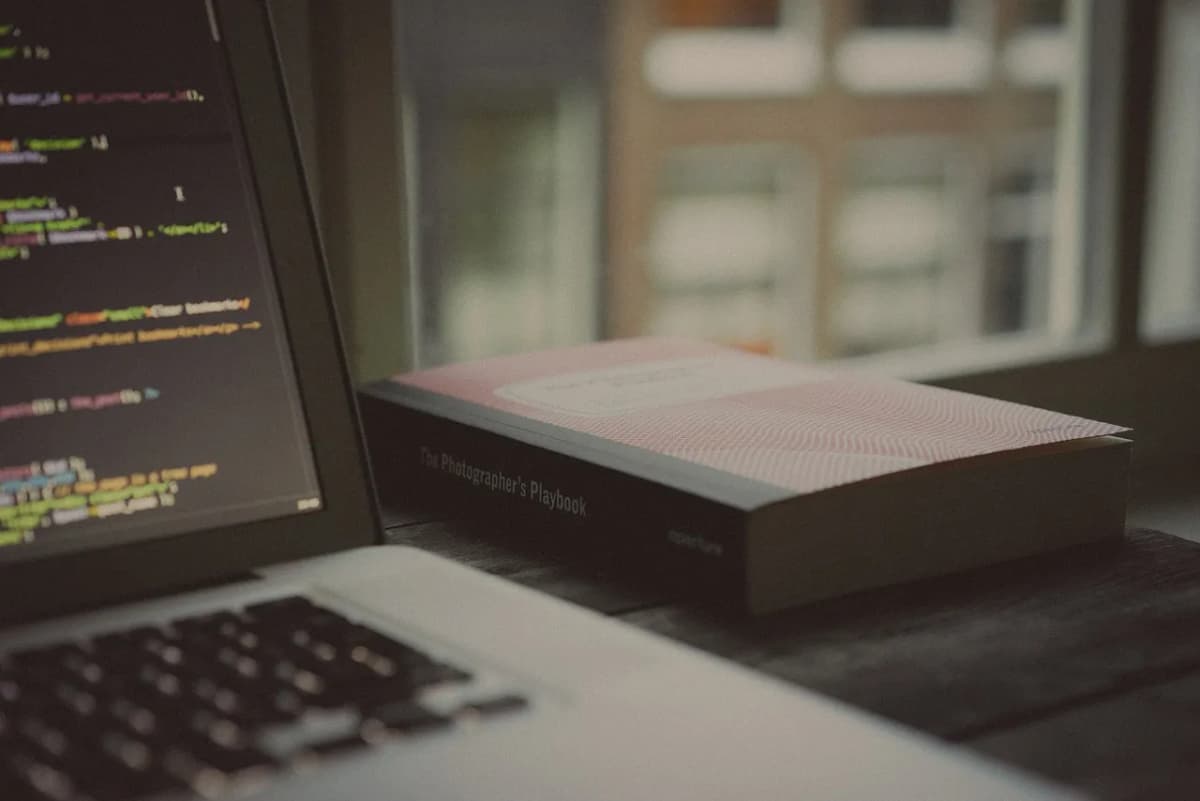 Laptop screen showing code beside a book on a wooden desk by a window