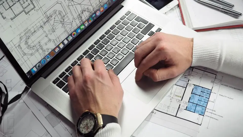 Hands typing on a laptop at a desk covered with printed plans, as someone checks an online job application