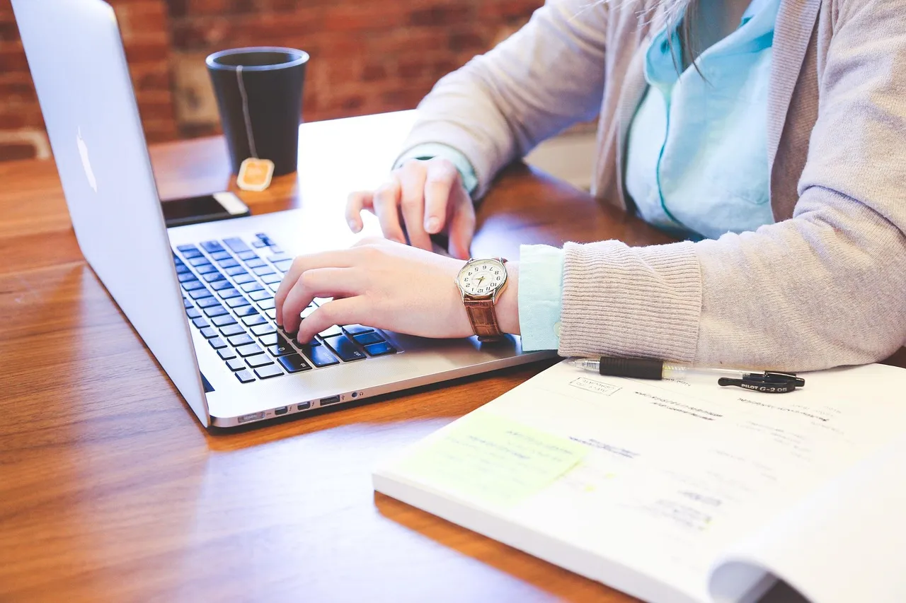 Hands typing on a laptop at a wooden desk beside an open notebook