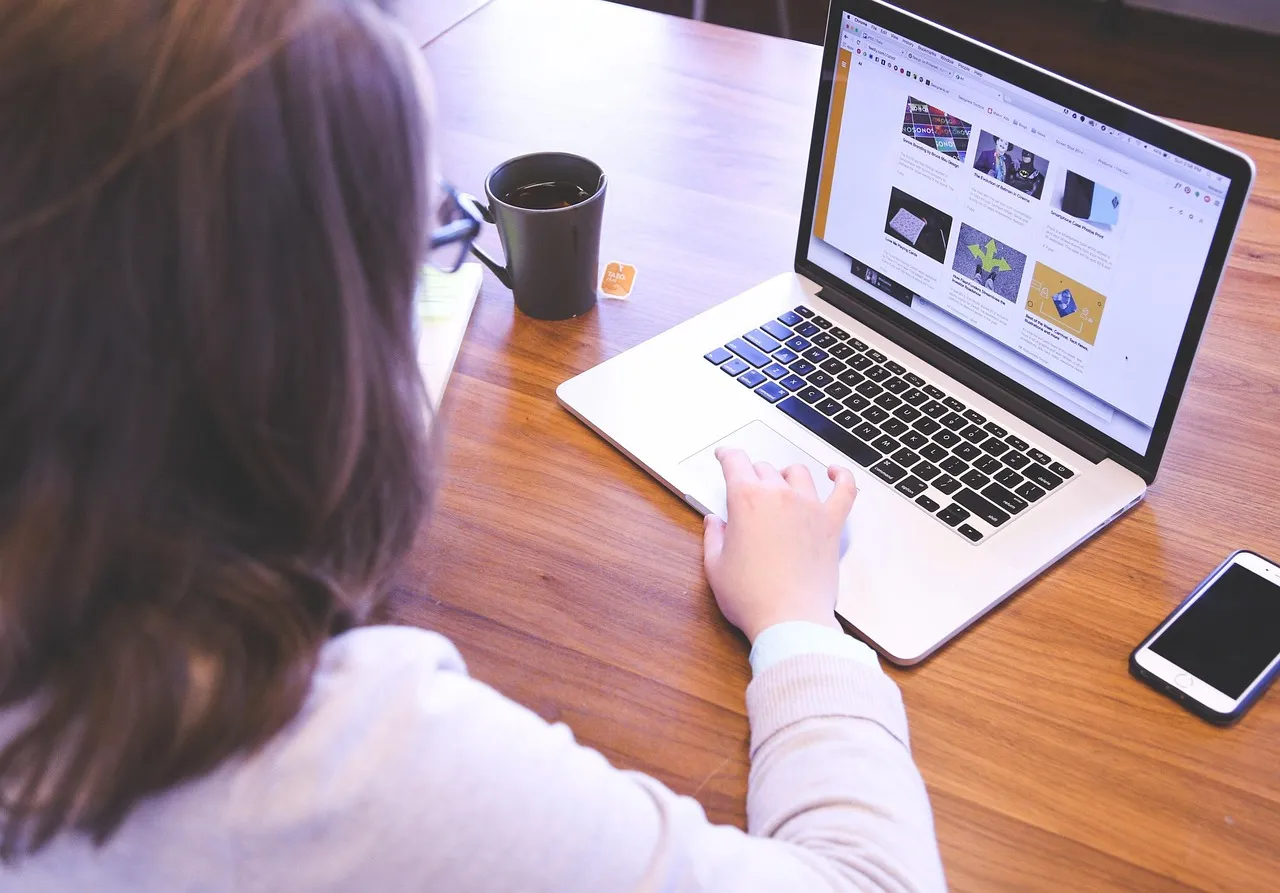 Woman seen from behind using a laptop showing a web page of images on a wooden desk with a mug and a phone