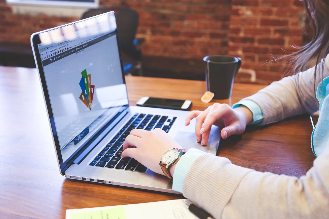 Person typing on a laptop showing a colourful design graphic at a wooden table with a phone and coffee cup