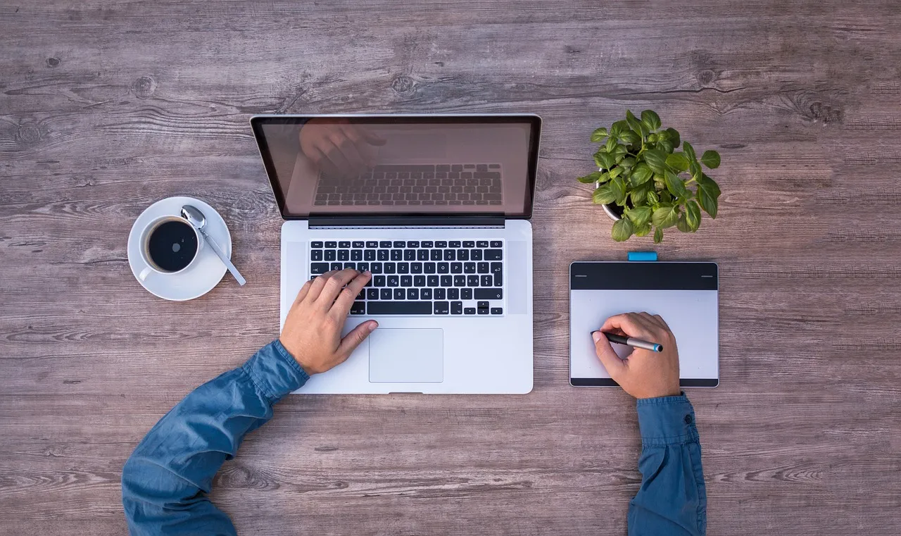 Overhead view of hands at a laptop and tablet on a wooden desk with coffee and a plant