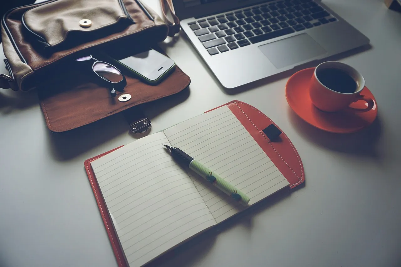 Open lined notebook with a pen beside a laptop, a red coffee cup and a leather bag on a desk