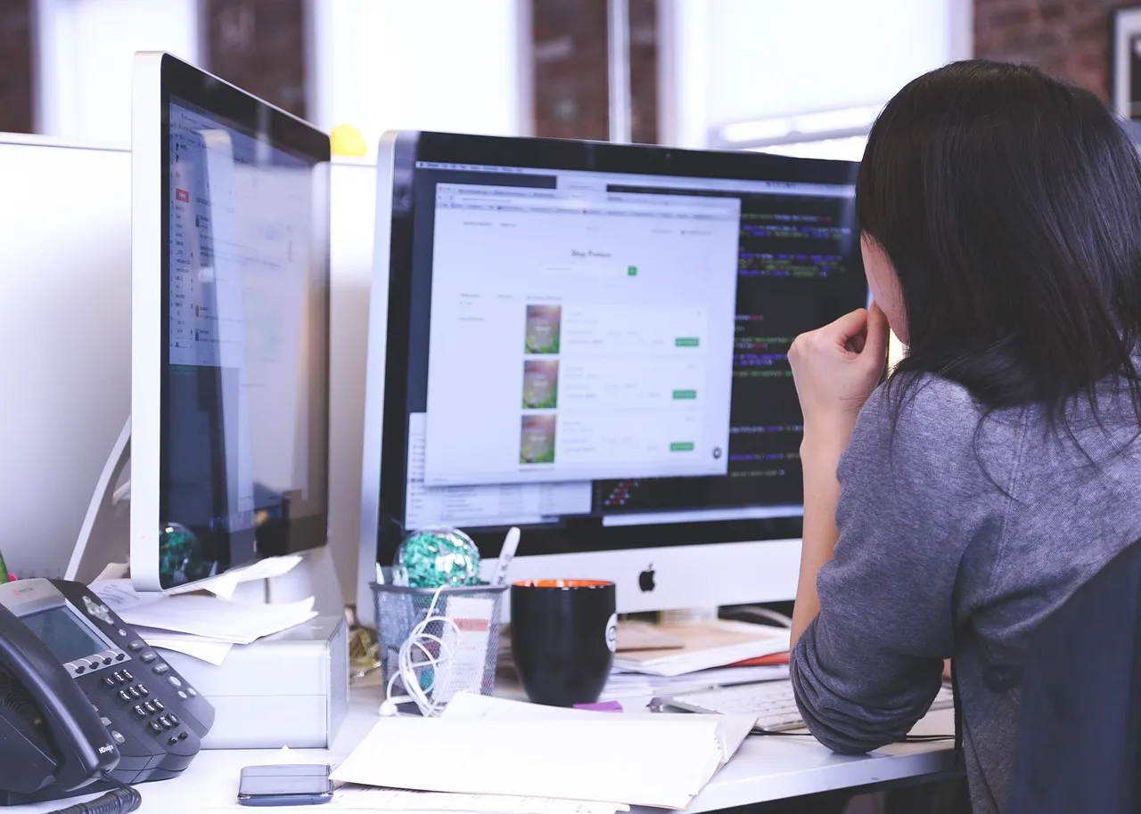 Woman working at a desk with two monitors showing a web page layout and code
