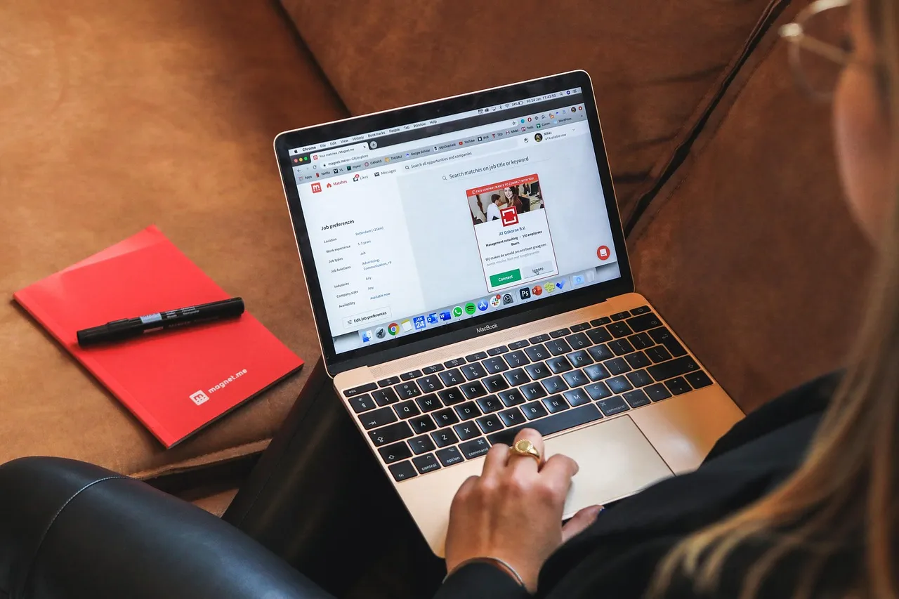 Woman browsing a website on a laptop on a sofa, next to a red notebook