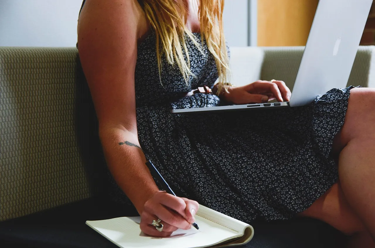 Woman on a sofa with a laptop on her lap writing notes in a notebook