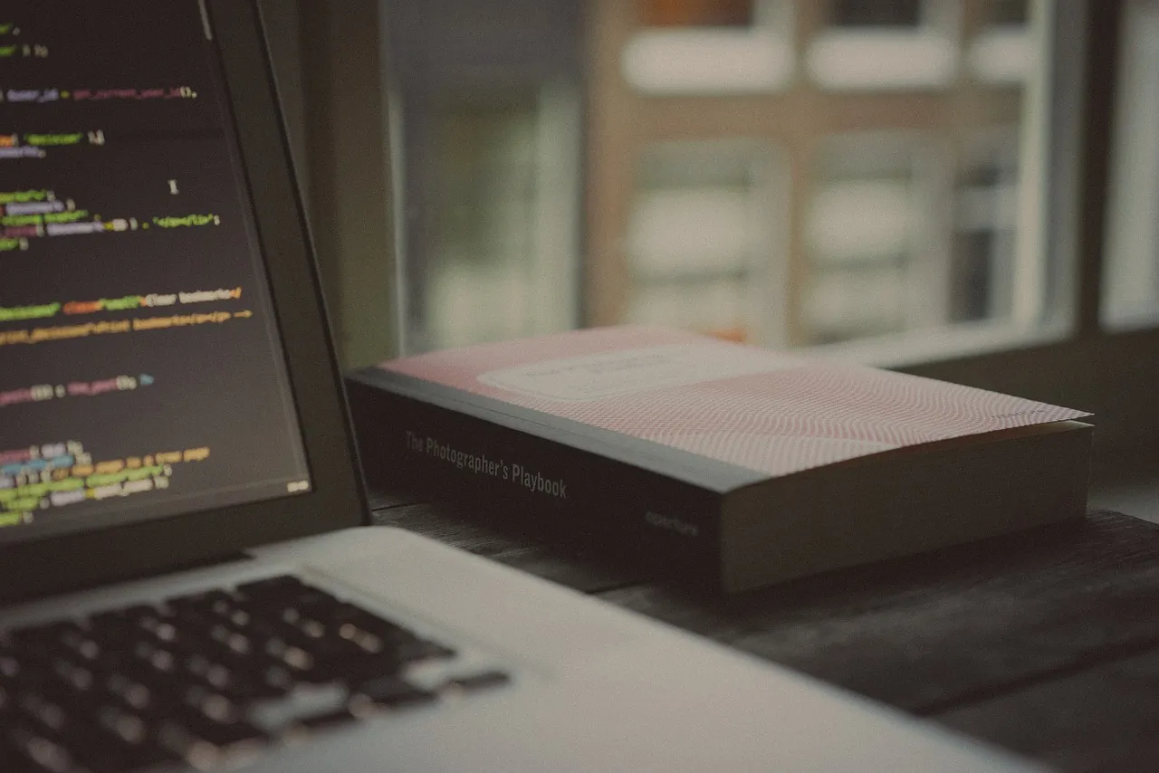 Laptop screen showing code beside a book on a wooden desk by a window