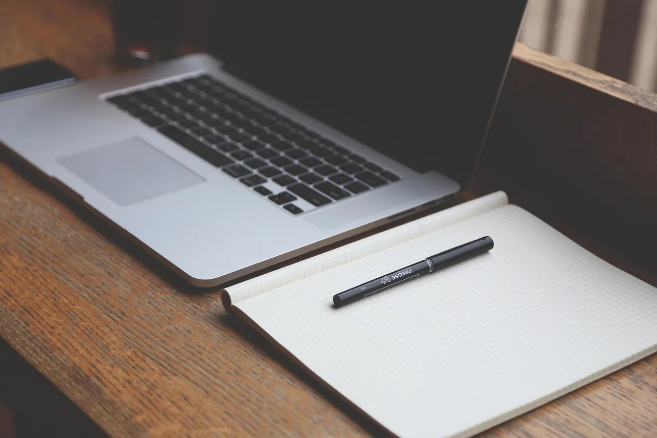Laptop on a wooden desk beside an open notebook and a black pen
