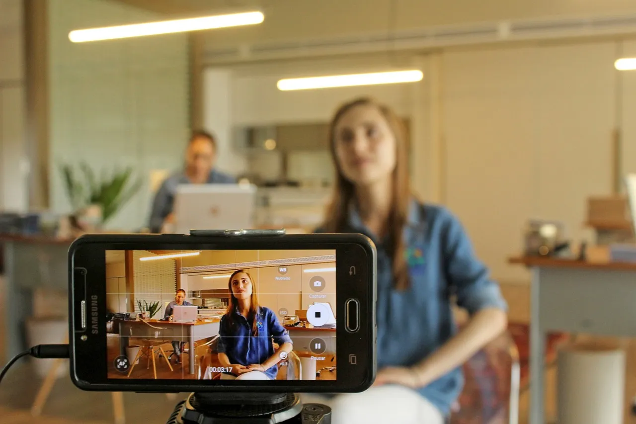 Smartphone on a tripod recording a woman in a denim shirt talking to camera in an office