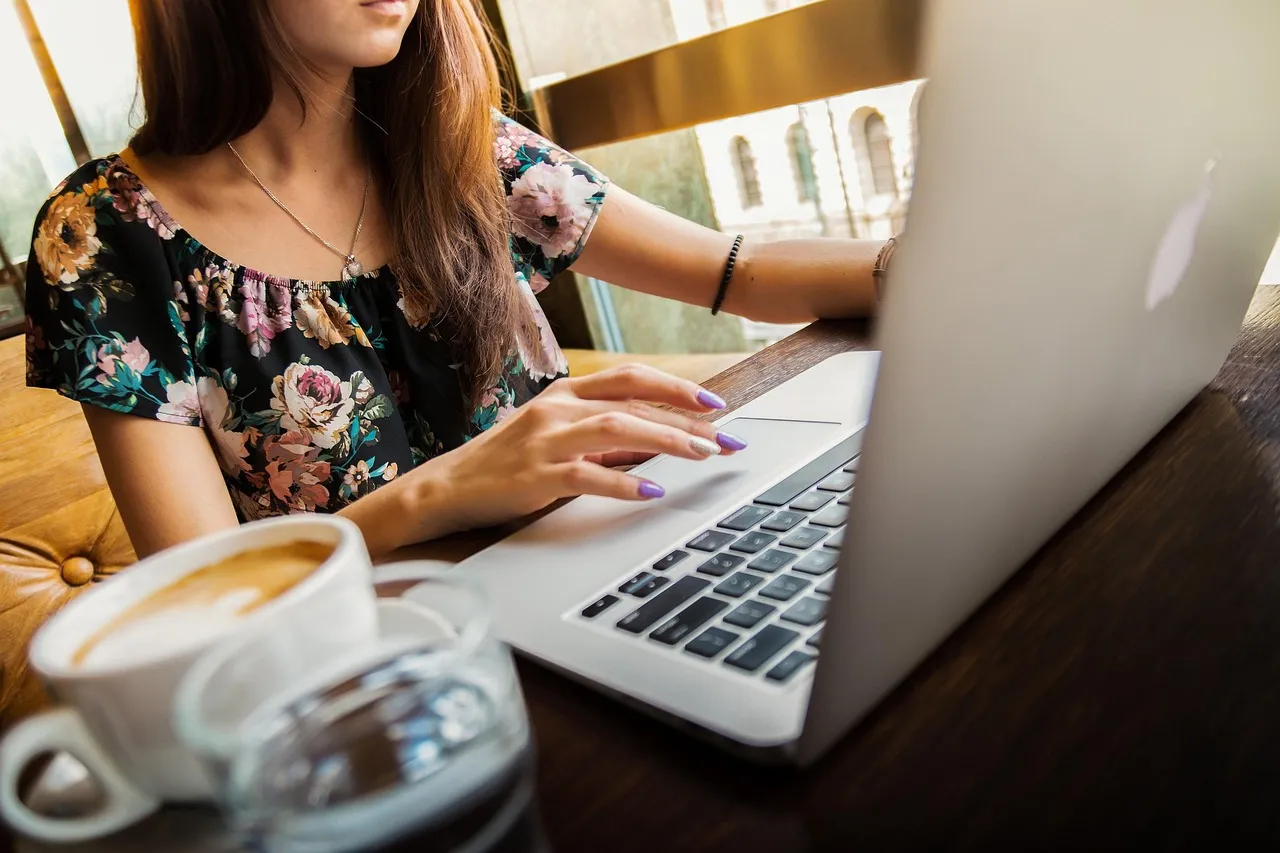 Woman in a floral top typing on a laptop at a cafe table next to a cup of coffee