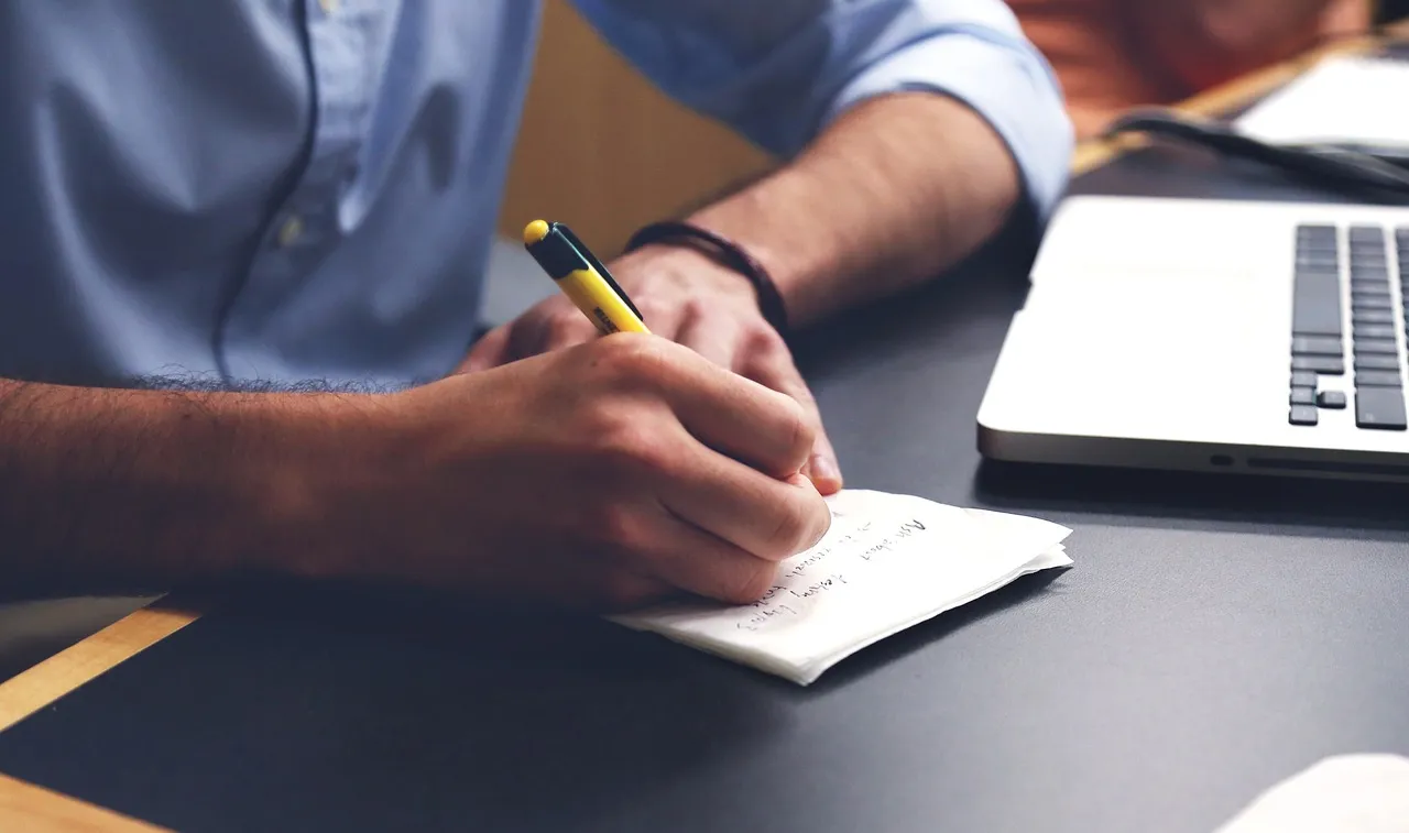 A man in a blue shirt writing notes with a pen beside an open laptop on a dark desk