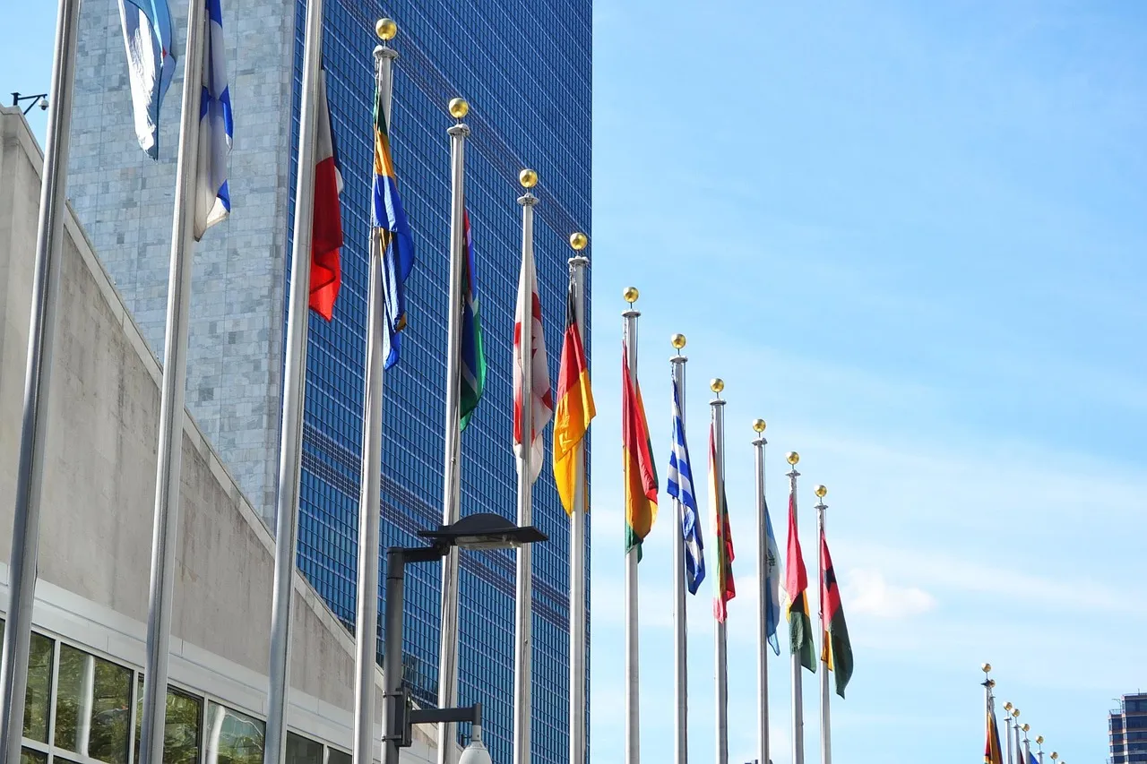 Row of national flags on tall poles in front of a glass skyscraper under a blue sky