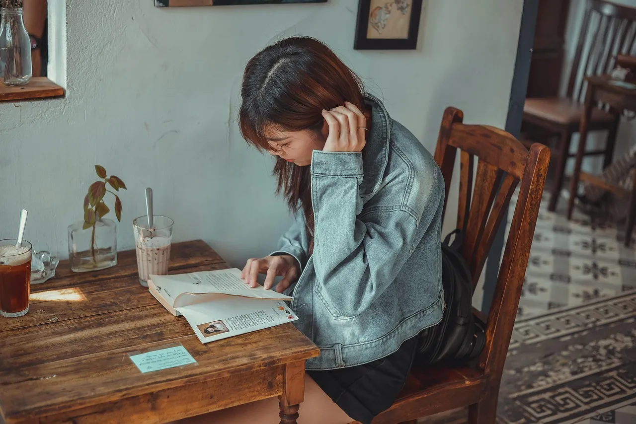 Young woman in a denim jacket reading a book at a wooden cafe table with drinks
