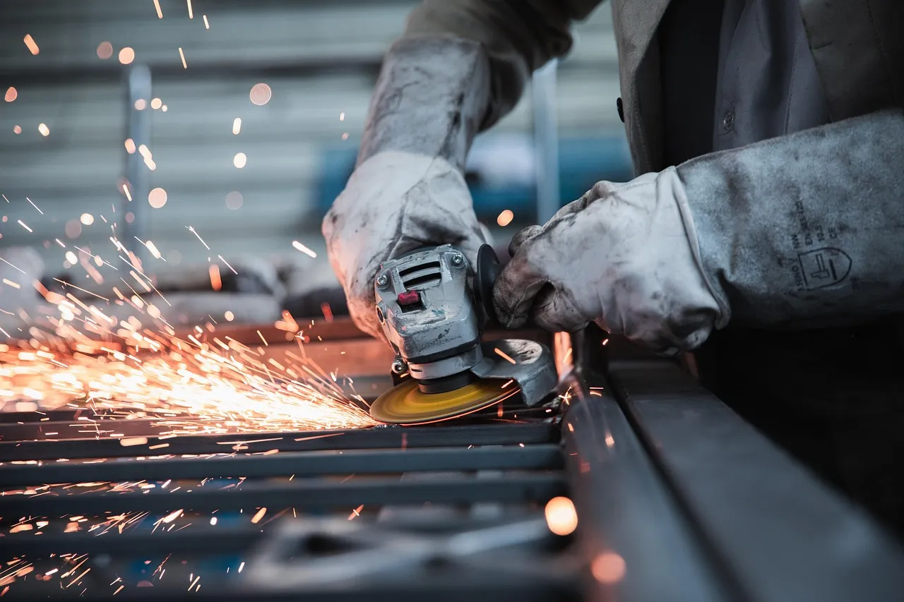 Worker in gloves using an angle grinder on metal, sending sparks across a factory bench