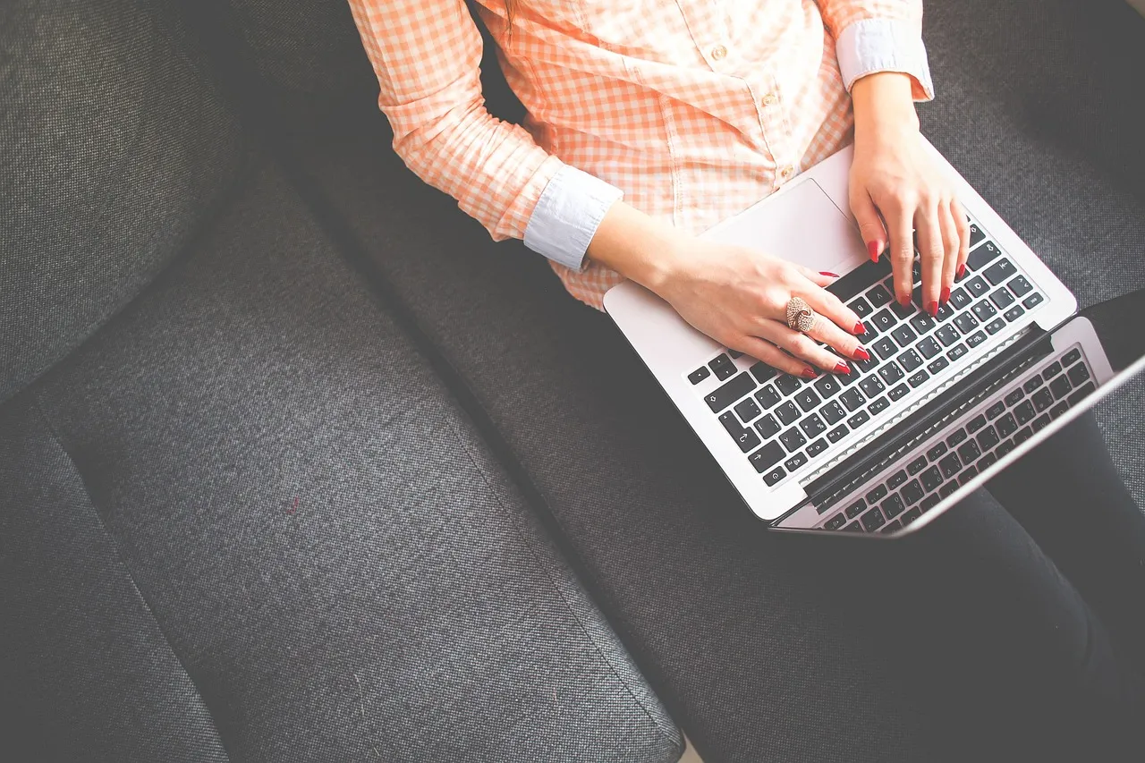 Person in a striped shirt typing on a laptop resting on a grey sofa