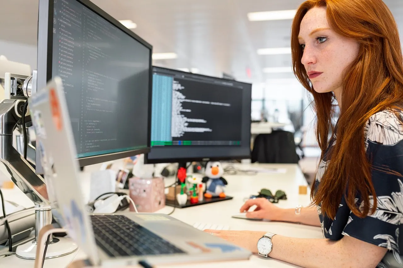 Woman with red hair working at a desk with two monitors showing code and a laptop