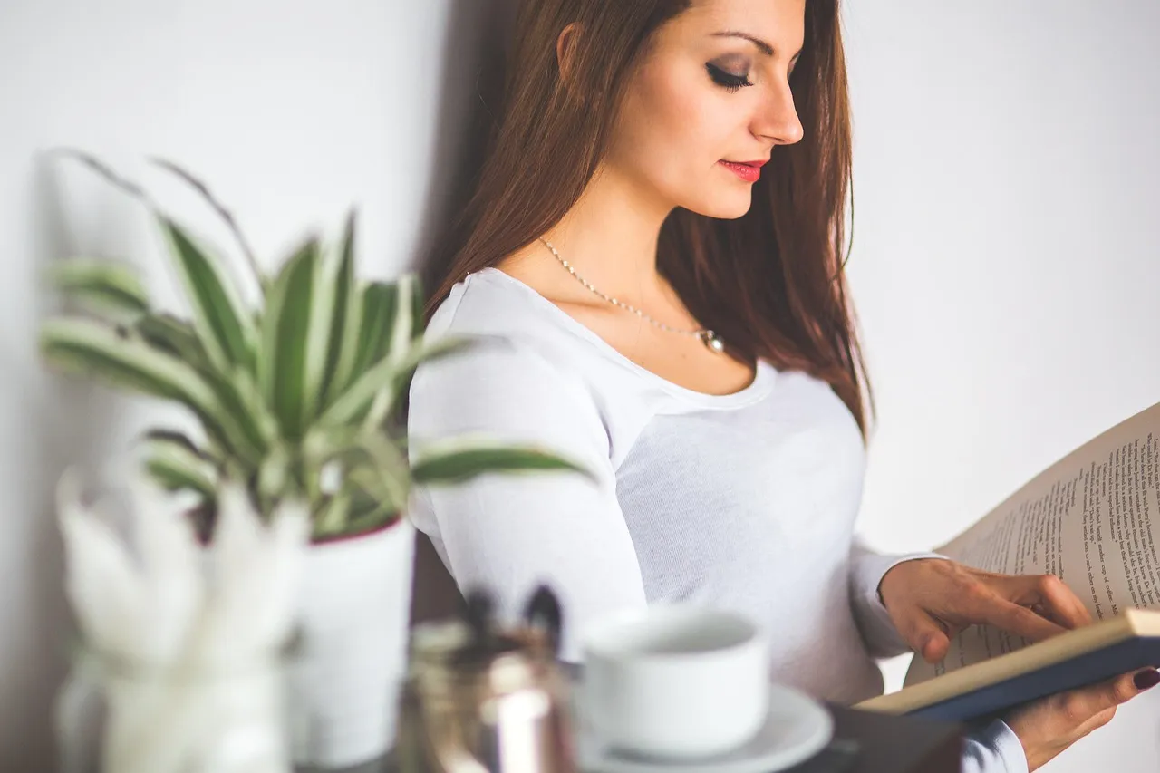 Woman in a white top reading an open book beside a coffee cup and a potted plant