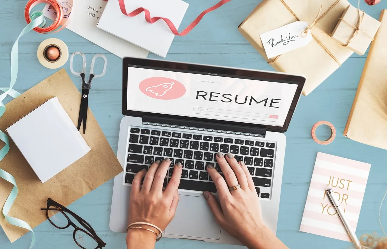 Hands typing on a laptop showing the word Resume, surrounded by craft supplies