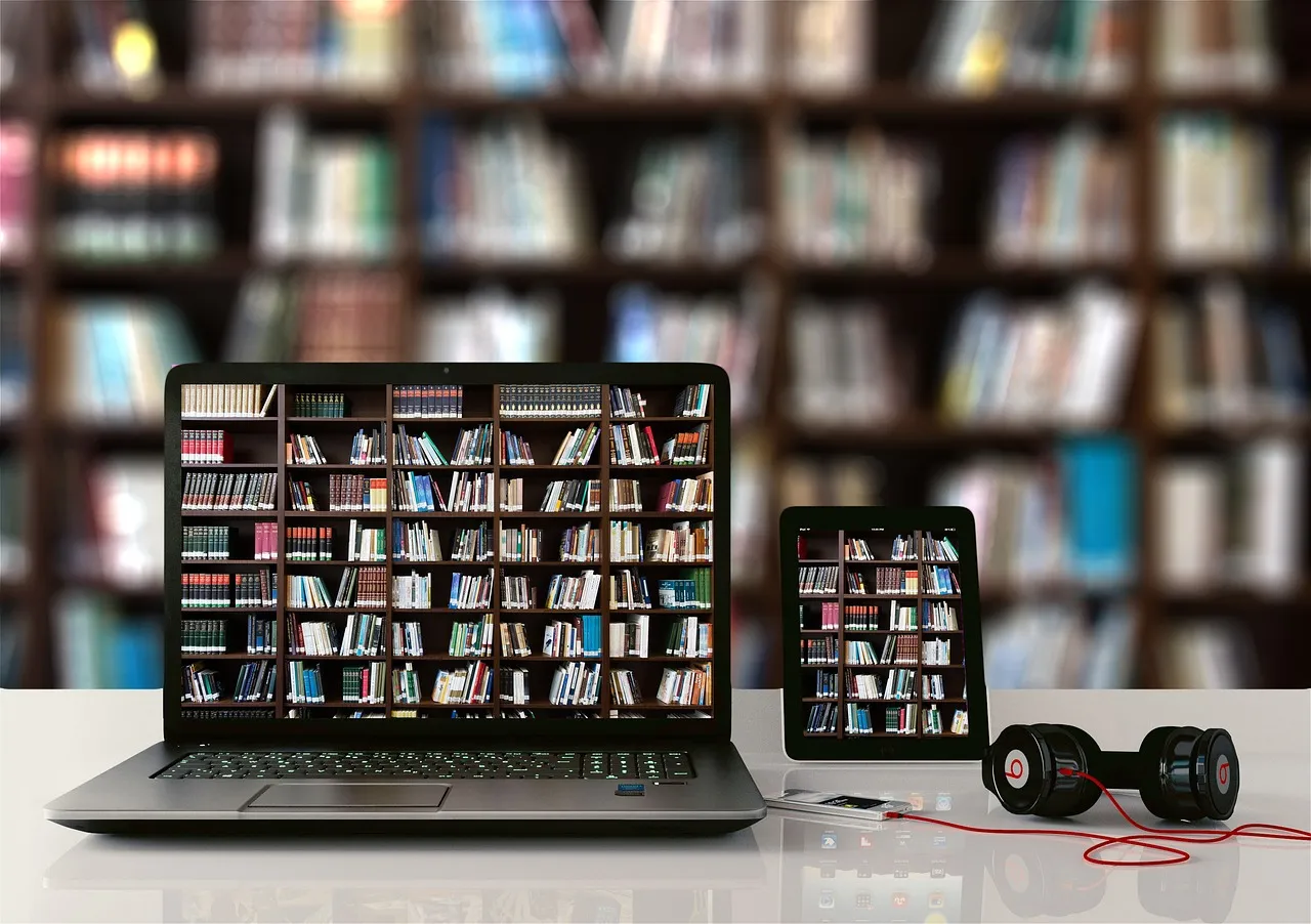 Laptop and tablet showing bookshelves on screen, with headphones on a desk in front of a library