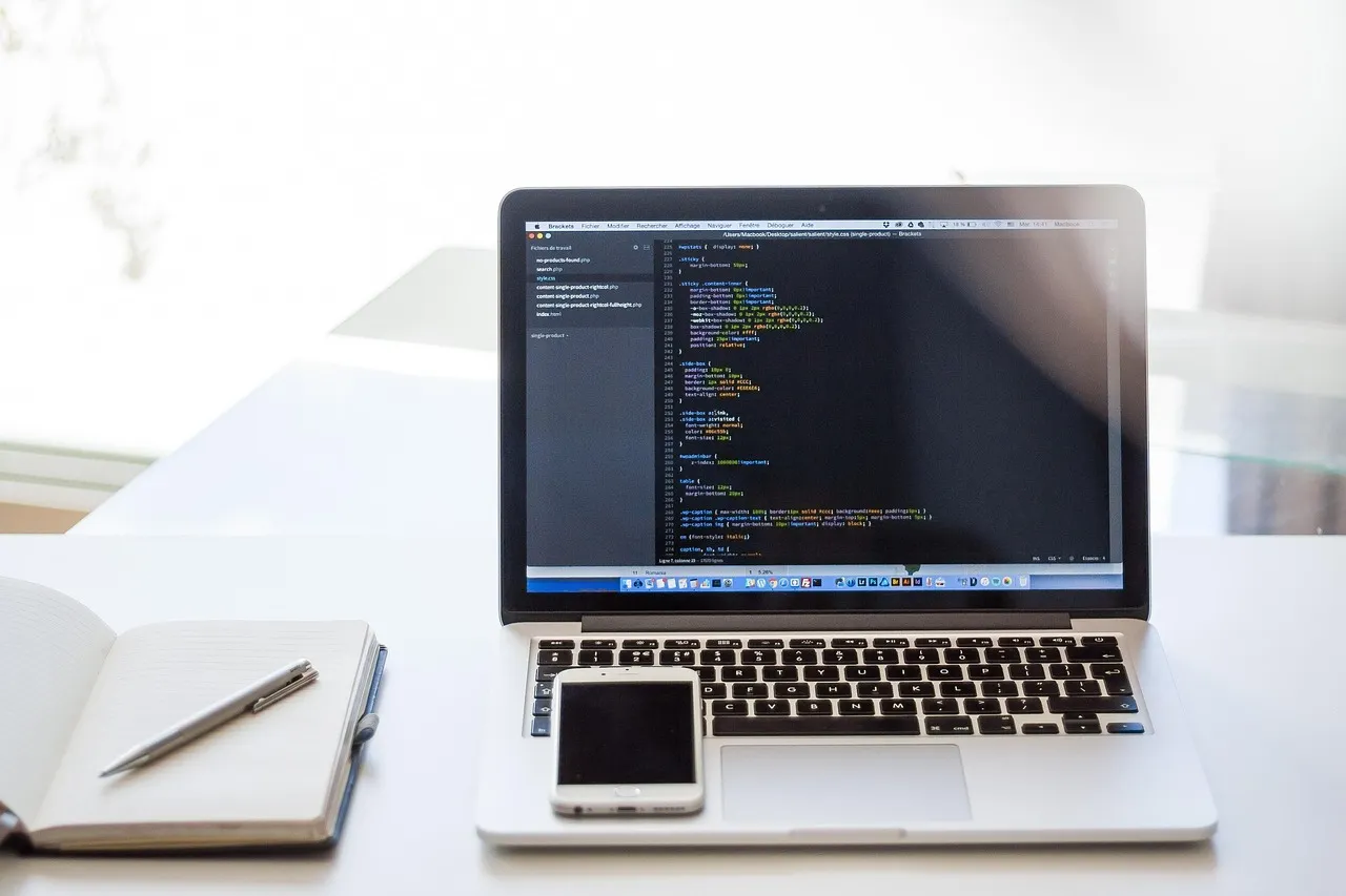 Open laptop showing lines of code on a white desk next to a smartphone, a notebook and a pen