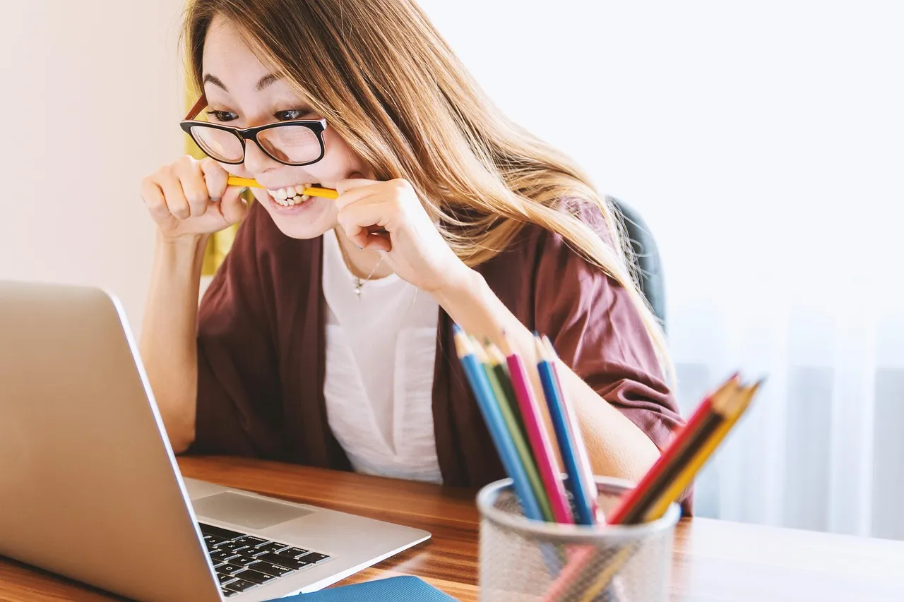 Young woman in glasses biting a pencil while staring at her laptop
