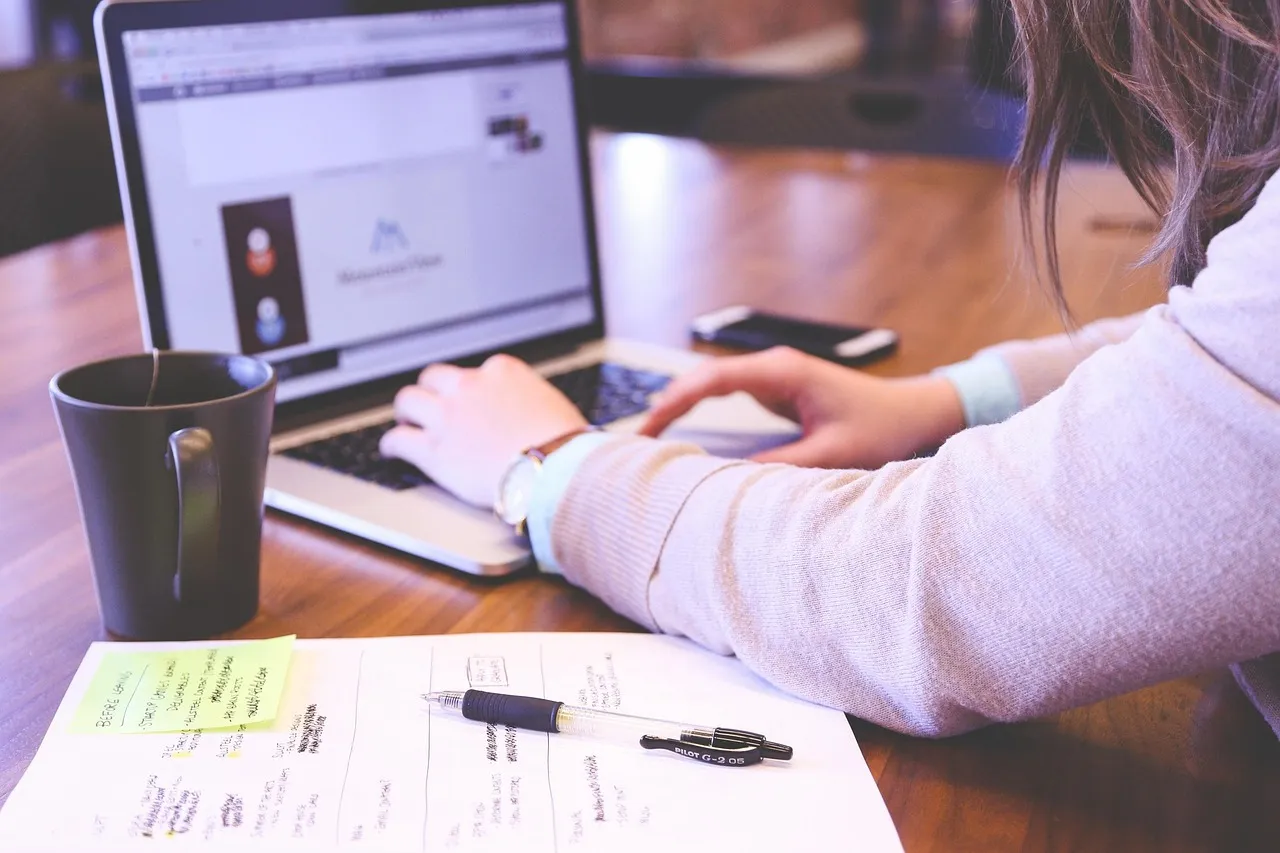Person typing on a laptop beside a coffee mug and handwritten notes